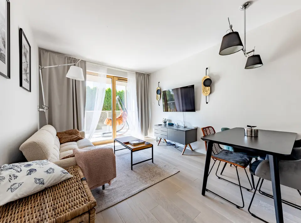 Kącik dzienny z wyjściem na balkon: jasna sofa obok niskiego drewnianego stolika kawowego, na przeciwnej ścianie TV zawieszony nad szarą komodą oraz czarny stół z krzesłami, za nimi przeszklone drzwi odsłaniające wiszące krzesło na zewnątrz.