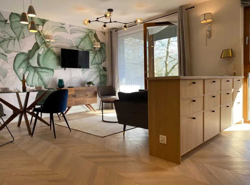 Salon z modnym muralem: ściana z tapetą w tropikalne liście, pod nią wiszący TV i konsola z drewna, drewniany stół jadalniany z krzesłami, otwarta przestrzeń dzienna z dużym oknem.