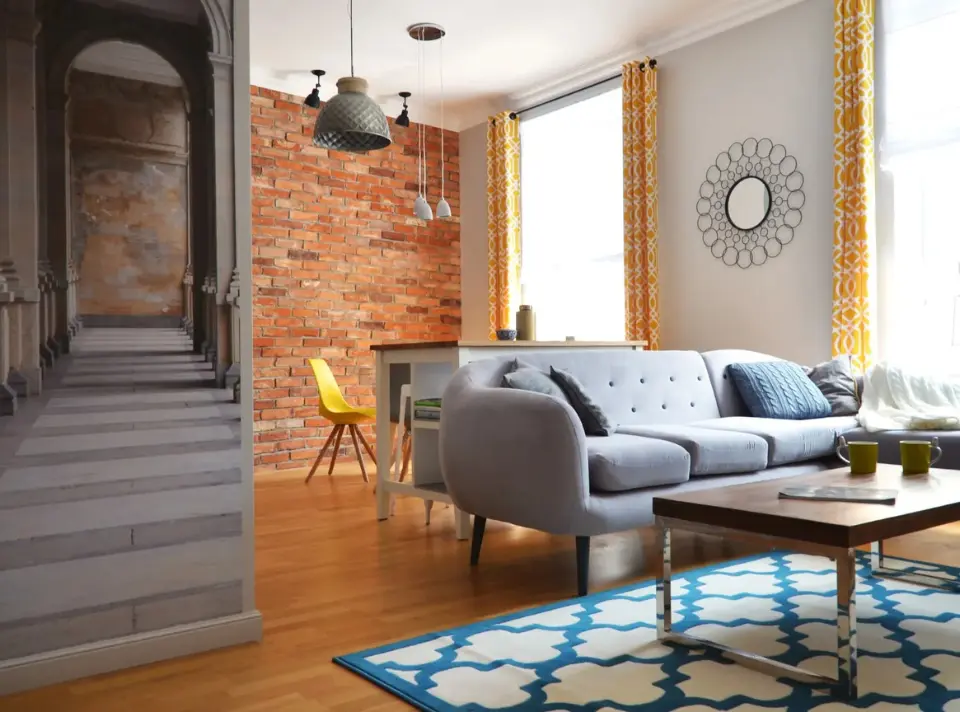Salon z akcentem ceglanej ściany: szara kanapa przed stolikiem kawowym, po lewej fragment ściany z architektoniczną fototapetą, w tle wyspa kuchenna i żółte zasłony przy dużym oknie.